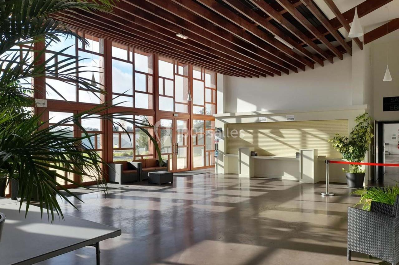 Hall lumineux avec de grandes baies vitrées, des plantes, un comptoir d'accueil et des sièges près des fenêtres.