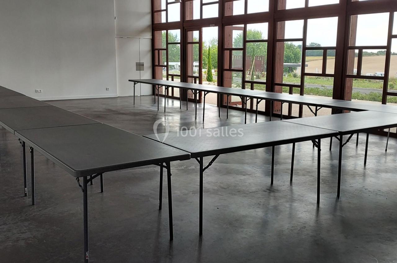 Salle lumineuse avec de grandes fenêtres, tables pliantes noires disposées en U sur un sol en béton.