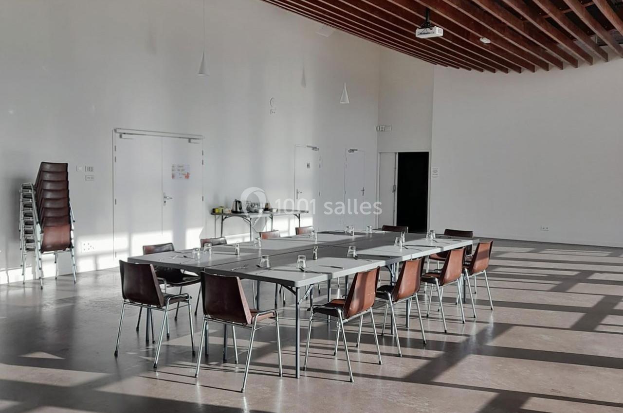 Salle lumineuse avec grandes baies vitrées, tables disposées en rectangle, chaises empilées et équipement de réunion.