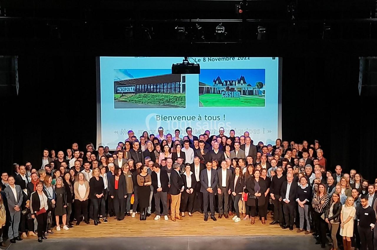 Photo de groupe sur une scène, avec un écran en arrière-plan affichant ’Bienvenue à tous !’ et des images de bâtiments.