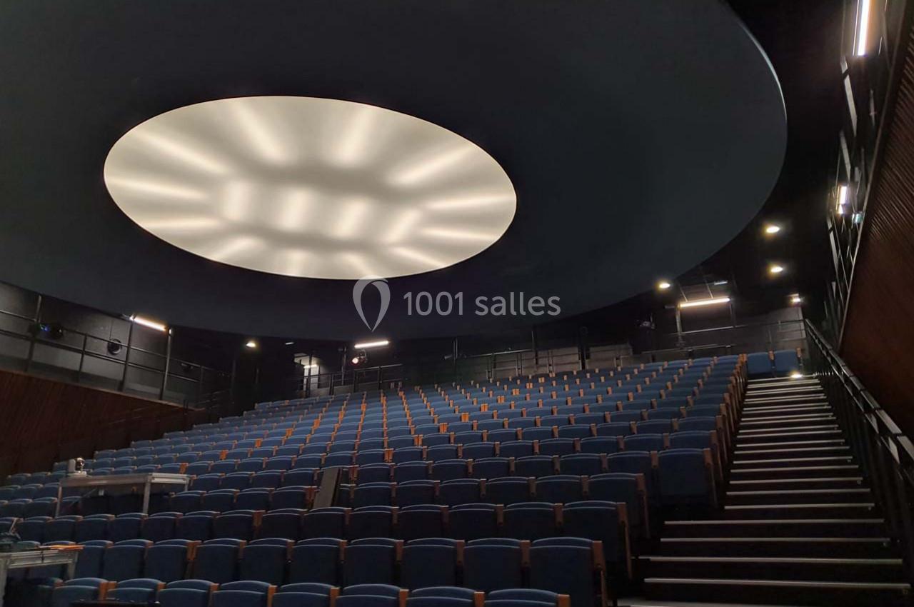 Salle de conférence vide avec des rangées de sièges bleus et un plafond circulaire éclairé au centre.