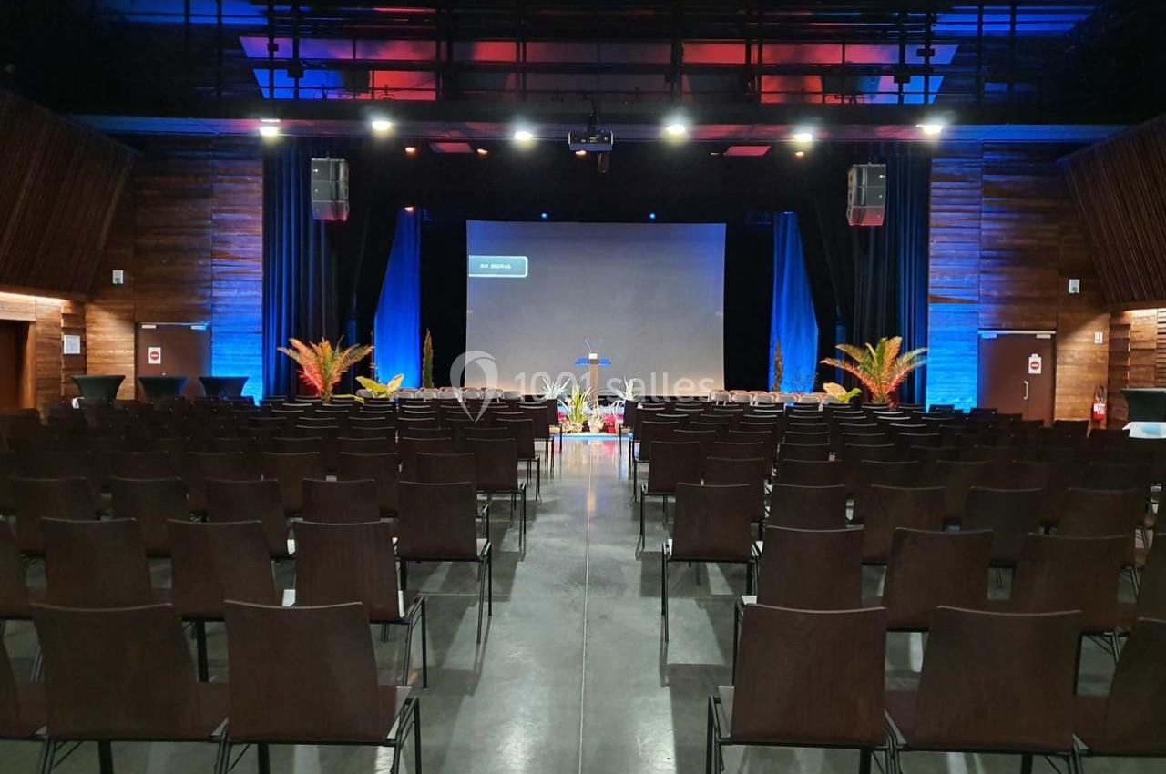Salle de conférence vide avec rangées de chaises, scène équipée d'un écran de projection et éclairage coloré.
