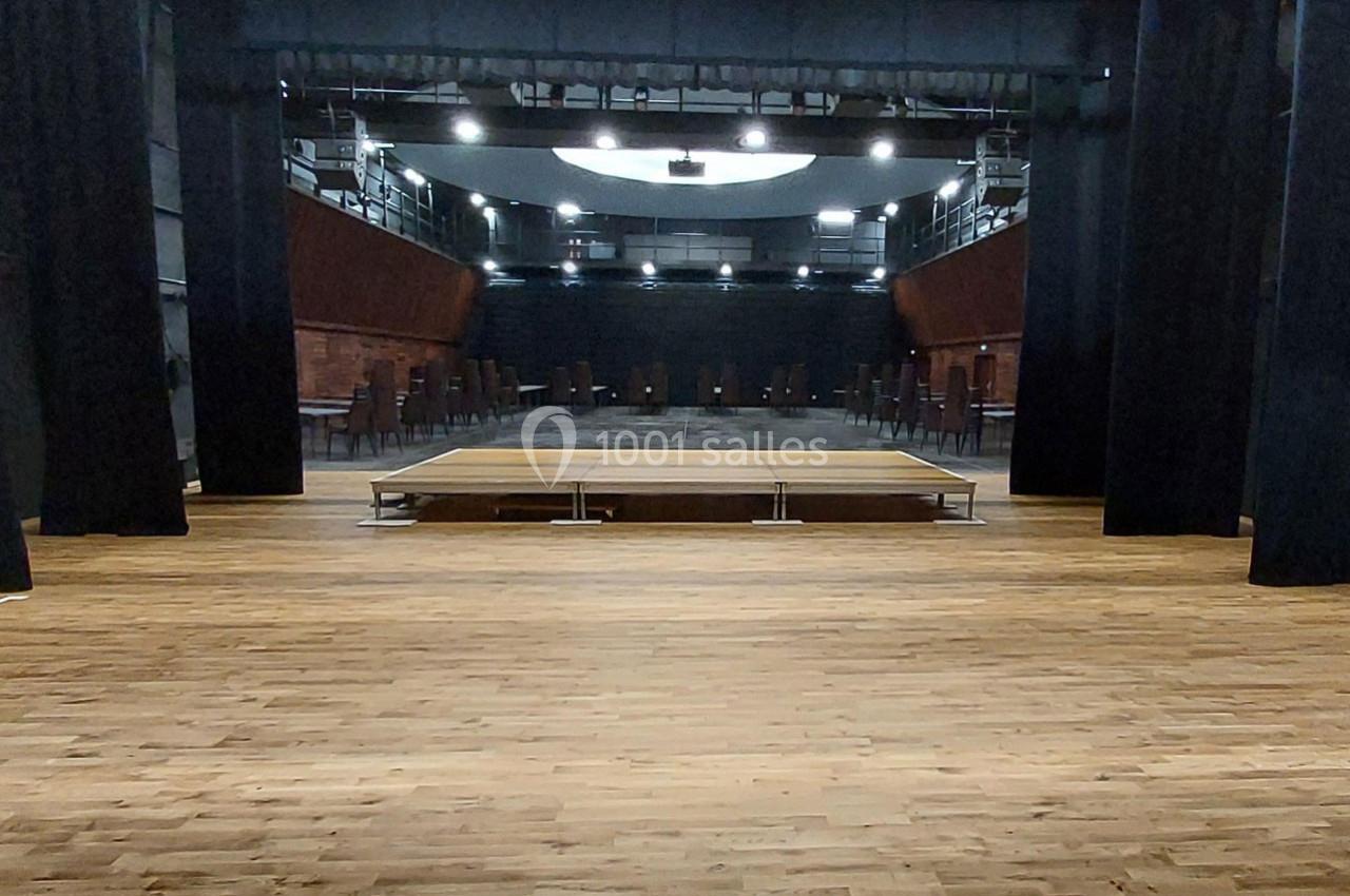 Salle de spectacle vide avec scène centrale en bois, gradins repliés et éclairage tamisé.
