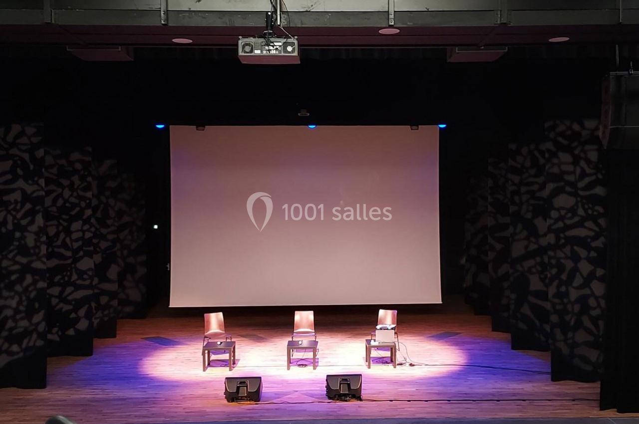 Trois chaises alignées sur une scène en bois avec un écran de projection blanc en arrière-plan.