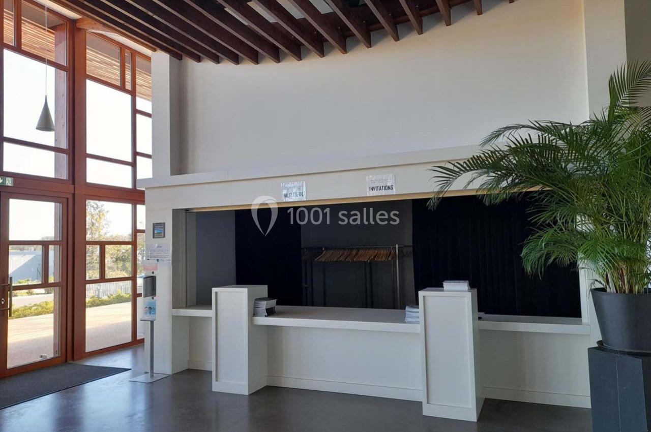 Accueil avec comptoir blanc, porte-manteaux en arrière-plan, grande baie vitrée et plante verte à droite.