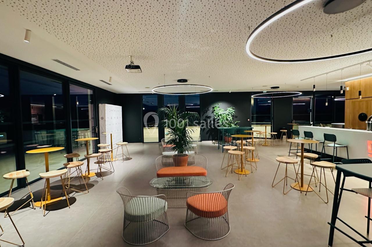 Salle moderne avec tables, chaises design, plantes et éclairage circulaire, vue sur un bar et de grandes fenêtres.