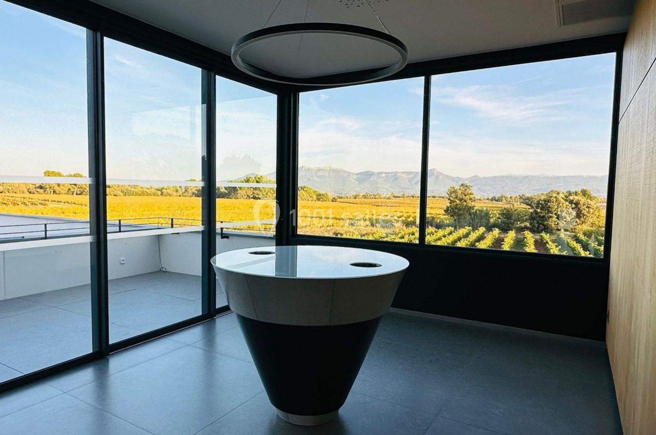 Salle lumineuse avec une table centrale noire et blanche, entourée de grandes baies vitrées offrant une vue sur des vignes…