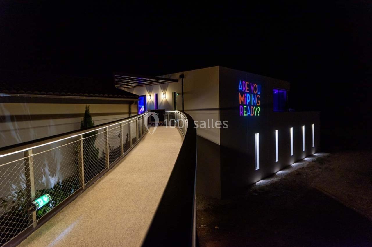 Passerelle éclairée menant à un bâtiment moderne avec des lumières colorées et un message lumineux sur la façade.