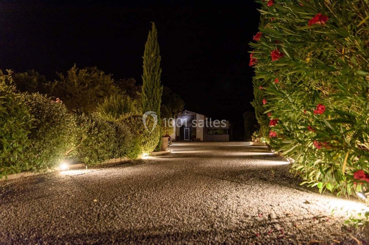 Allée de gravier éclairée par des lumières au sol, bordée de buissons et menant à une maison en pierre, de nuit.