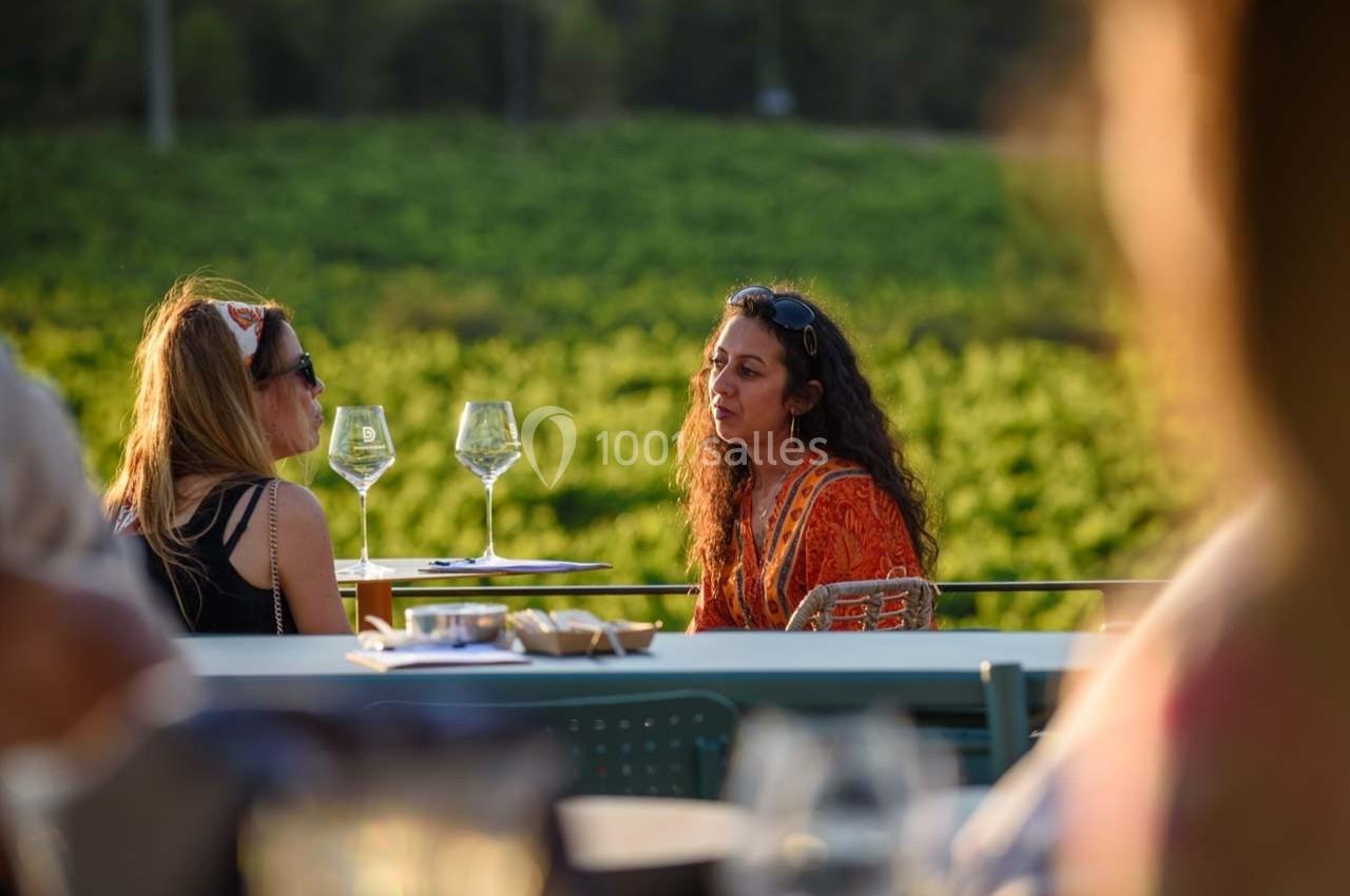 Deux femmes discutent à une table en extérieur, avec des verres de vin et un vignoble en arrière-plan.