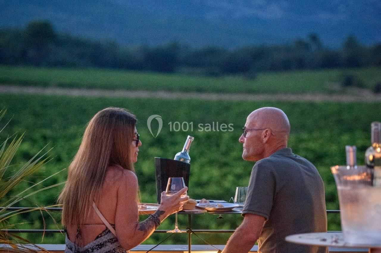 Un couple discute autour d'un verre de vin en terrasse au coucher du soleil, avec des montagnes en arrière-plan.