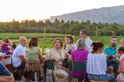 Des personnes assises à une table en extérieur, discutant et partageant un repas devant un paysage de vignobles au coucher…