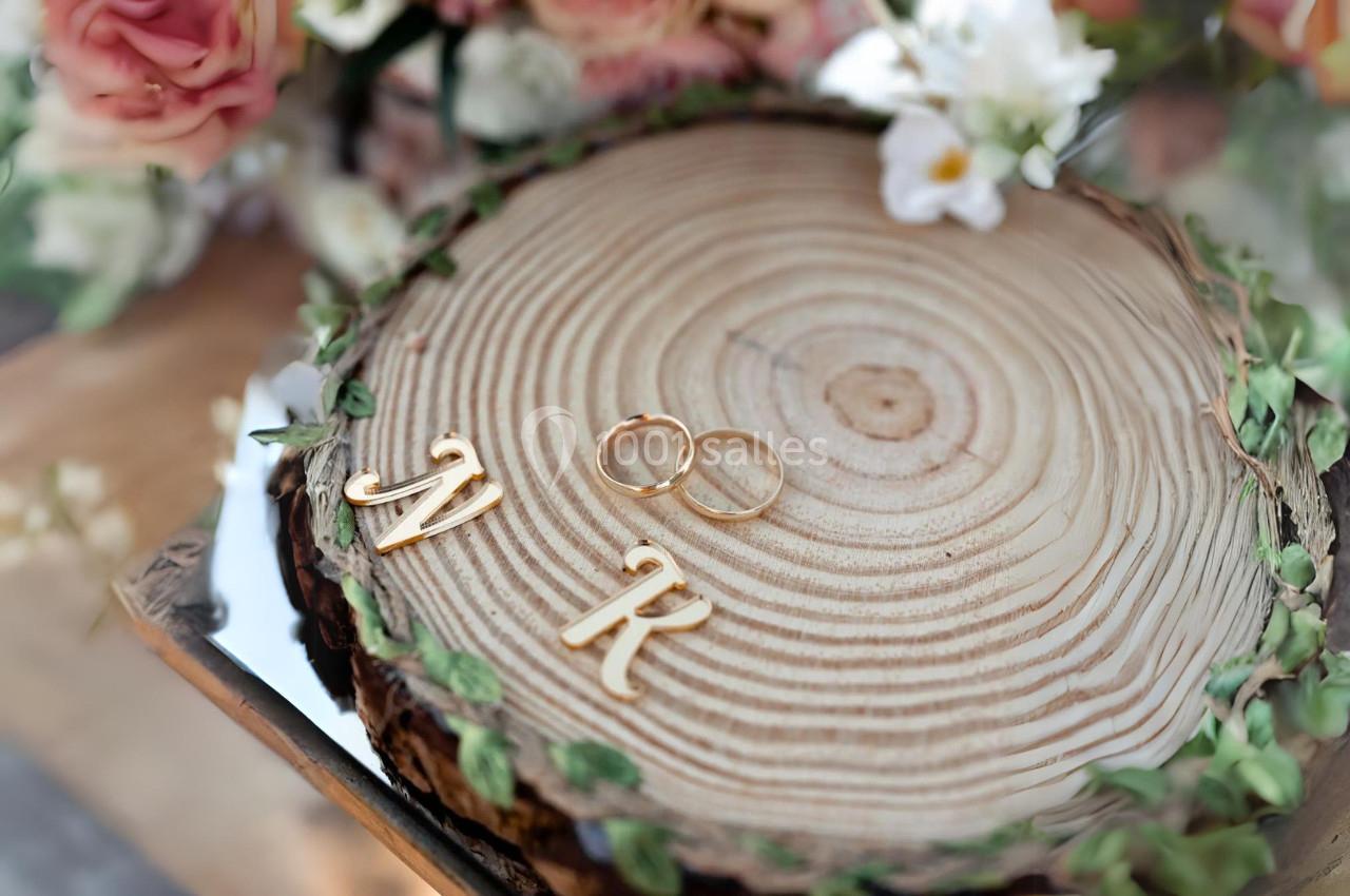 Deux alliances dorées posées sur une tranche de bois décorée de feuillage, avec les lettres ’M’ et ’R’ en métal doré.