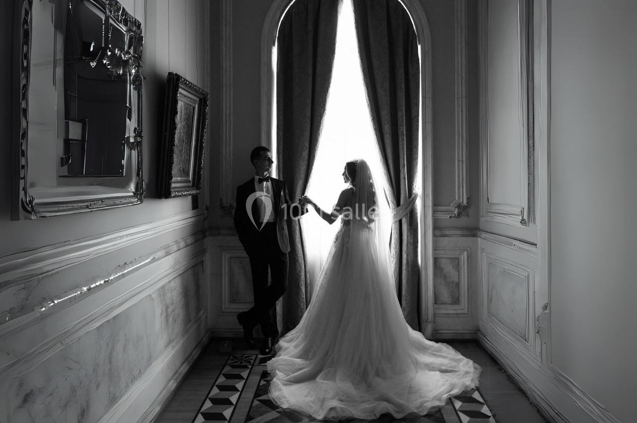 Un couple en tenue de mariage se tient près d'une grande fenêtre avec des rideaux, dans une pièce élégante.