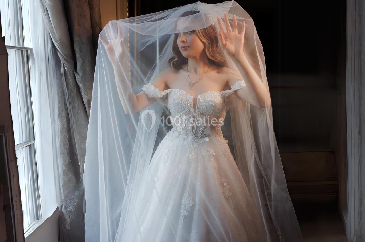 Femme en robe de mariée avec un voile, debout près d'une fenêtre, éclairée par une lumière douce.