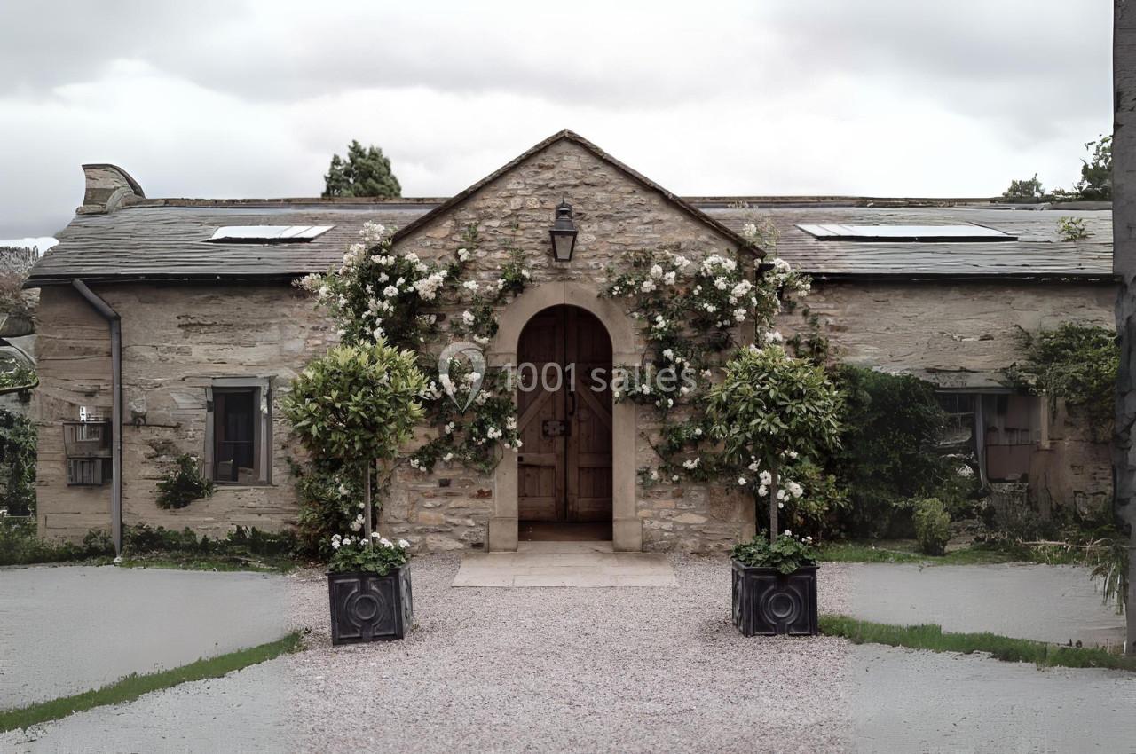 Façade en pierre d'une maison avec porte en bois, plantes grimpantes et deux grands pots fleuris devant l'entrée.