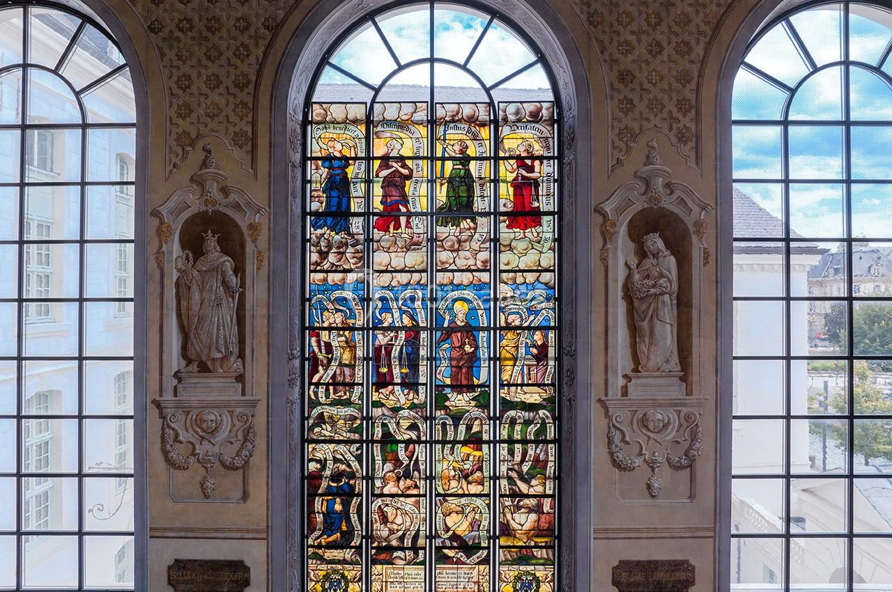 Vitrail coloré représentant des scènes religieuses, encadré par deux statues et des fenêtres laissant entrer la lumière.