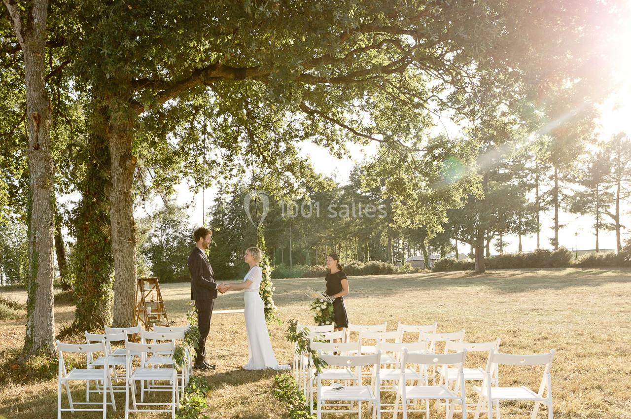 Un couple échange ses vœux sous un grand arbre dans un cadre champêtre, avec des chaises blanches disposées en allée.
