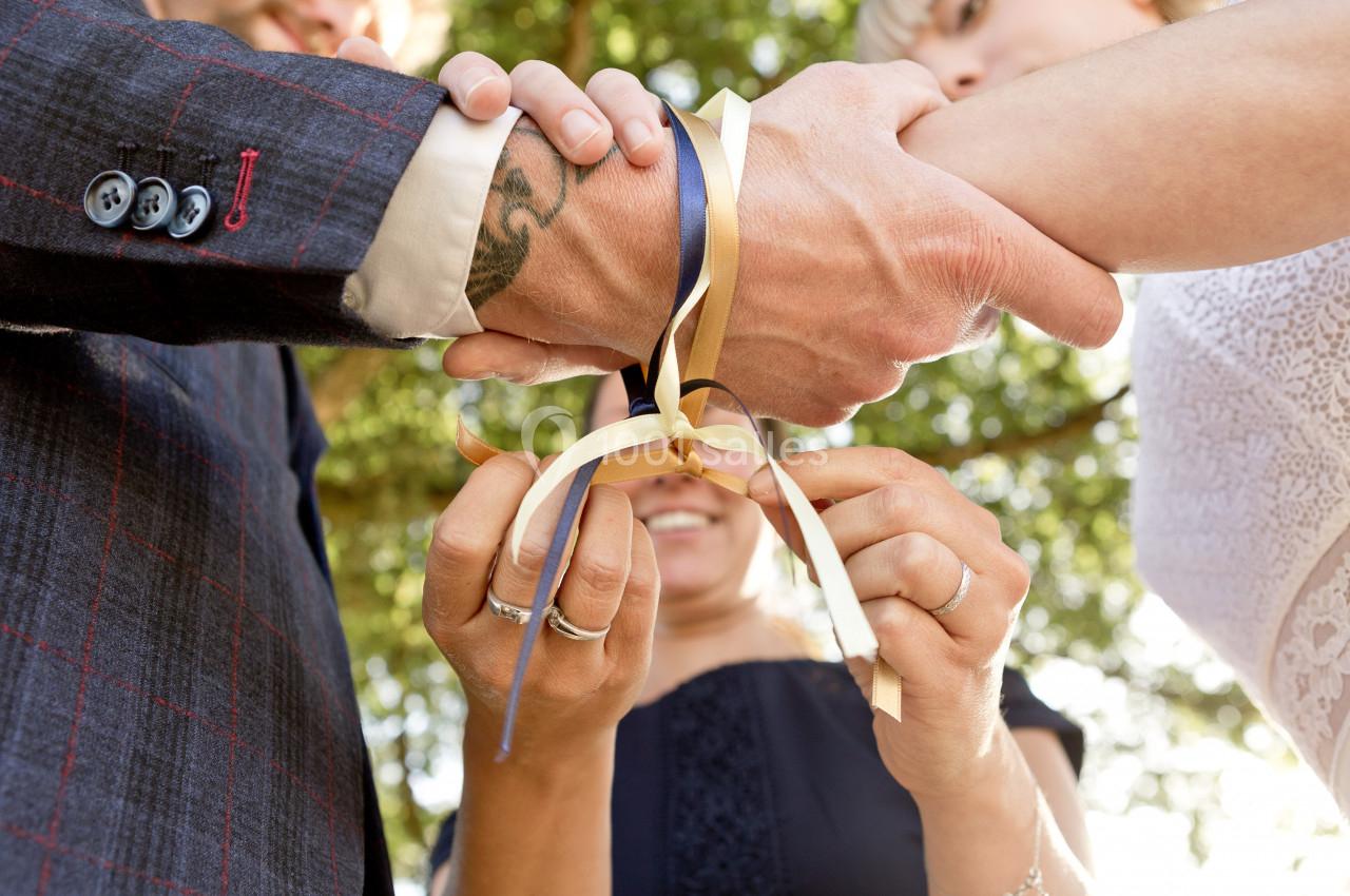 Deux mains liées par des rubans colorés, tenus par une troisième personne lors d'une cérémonie en extérieur.
