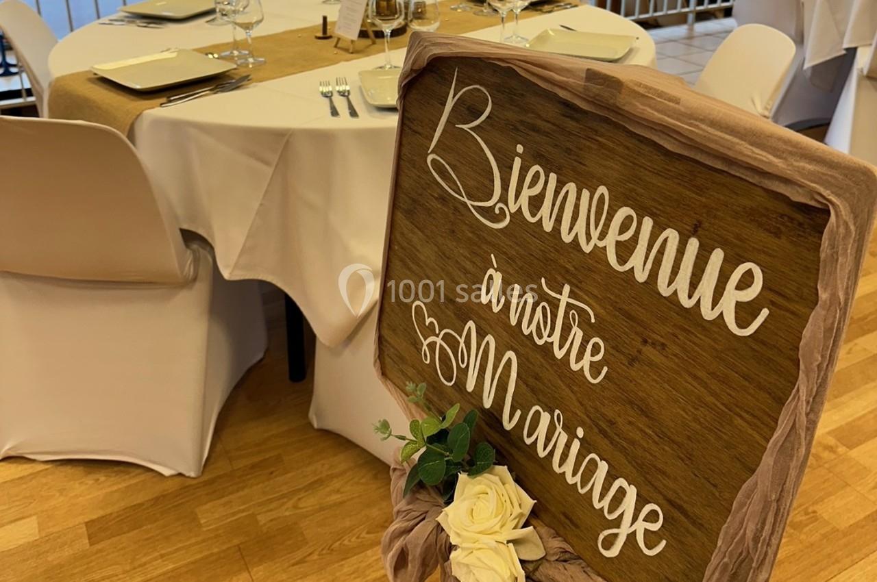 Panneau en bois avec l'inscription ’Bienvenue à notre mariage’, décoré de roses blanches, devant une table dressée.