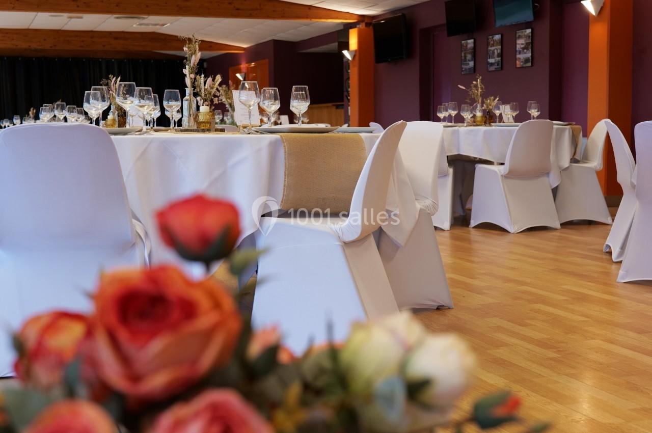 Salle décorée pour un événement avec des tables dressées, nappes blanches et bouquets de fleurs en premier plan.