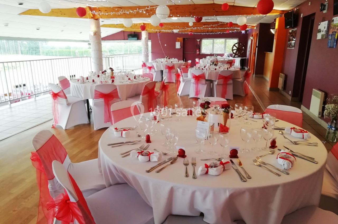 Salle décorée pour un événement avec des tables rondes dressées, nappes blanches, chaises ornées de rubans rouges.