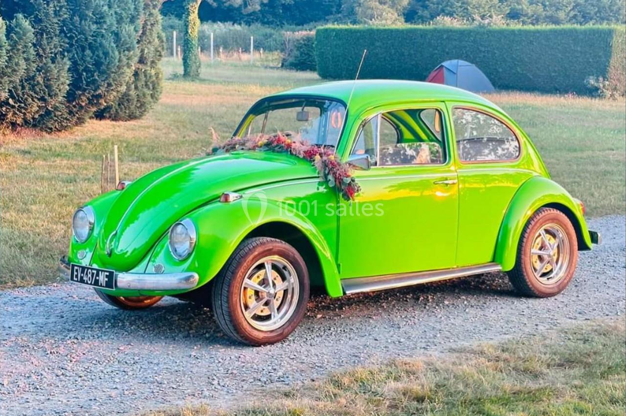 Voiture ancienne verte décorée de fleurs, stationnée sur un chemin gravillonné près d'une tente et d'arbres.