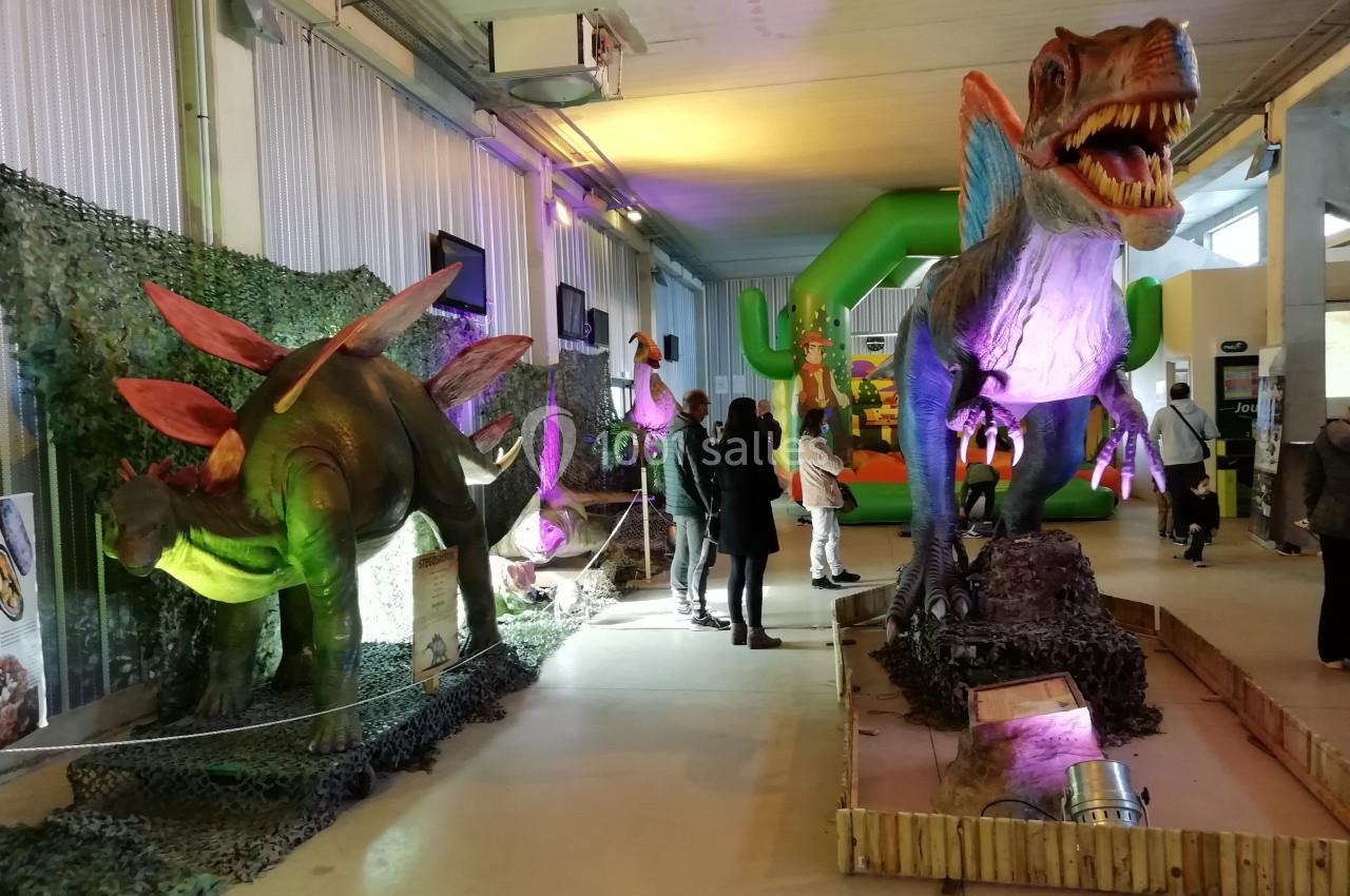 Exposition intérieure avec des dinosaures animés grandeur nature, éclairages colorés et visiteurs observant les modèles.