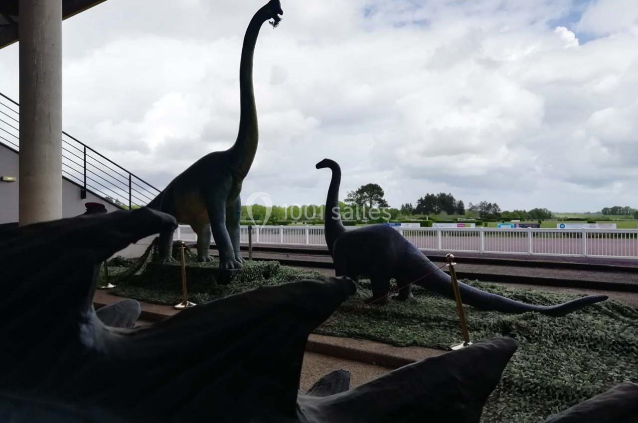 Statues de dinosaures herbivores sur un espace extérieur, avec un ciel nuageux et une barrière en arrière-plan.