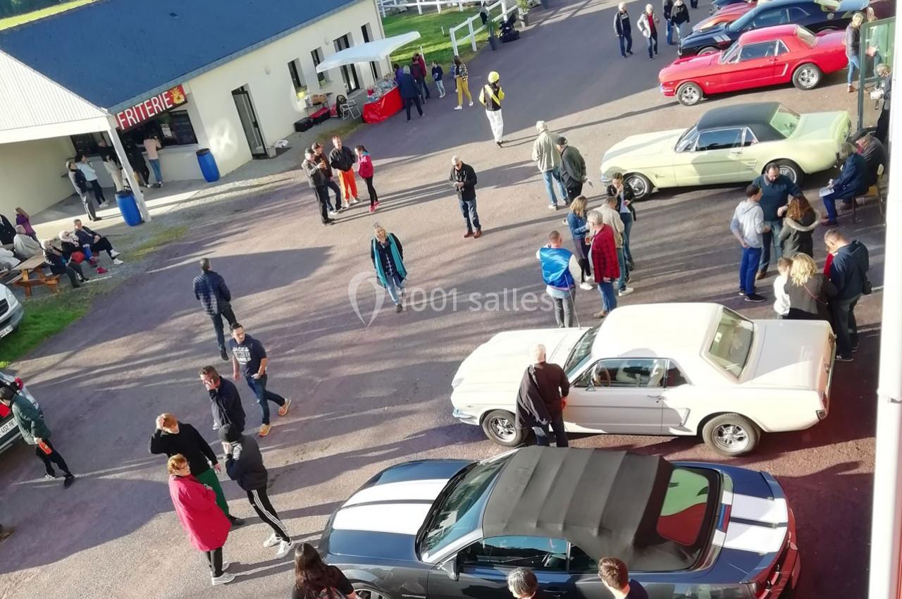 Rassemblement de personnes autour de voitures anciennes et modernes dans un parking près d'un bâtiment.