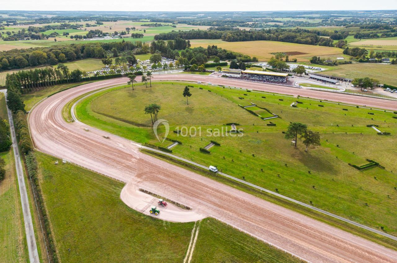 Vue aérienne d'un hippodrome entouré de champs, avec des pistes, des tribunes et des véhicules stationnés.