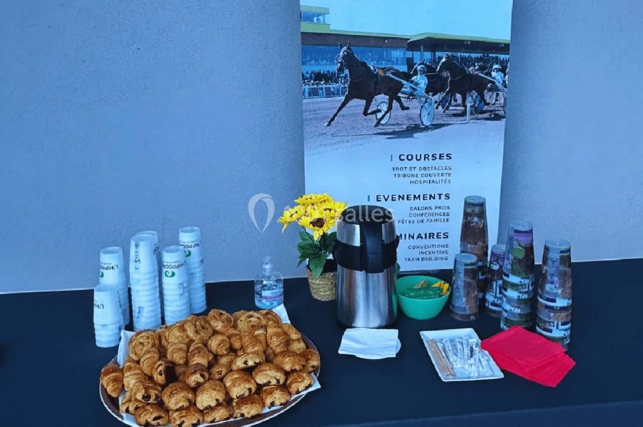 Table avec viennoiseries, gobelets, cafetière et dépliants devant une affiche de courses hippiques.