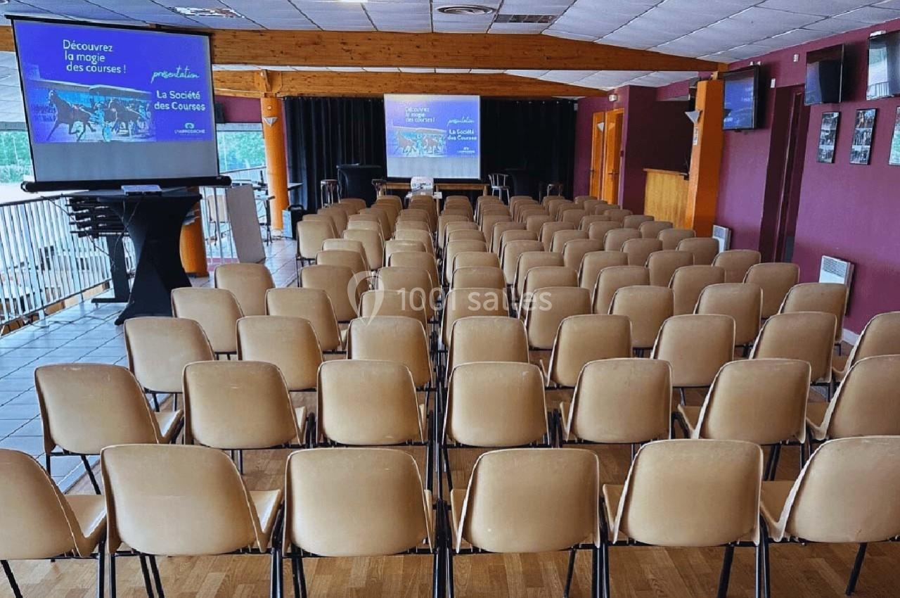 Salle de conférence avec rangées de chaises vides face à un écran de projection et une estrade.