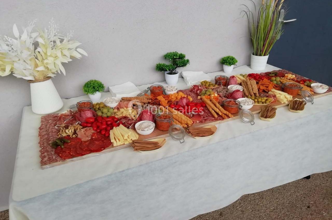 Table garnie de charcuteries, fromages, fruits, légumes, pains et condiments, décorée de plantes et vases blancs.