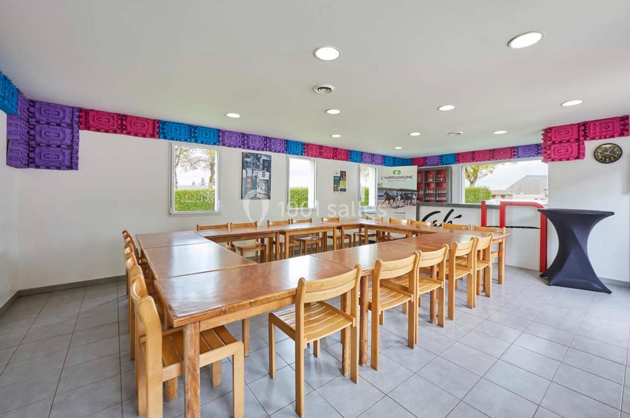 Salle lumineuse avec tables en bois disposées en U, chaises assorties, murs décorés et fenêtres donnant sur l'extérieur.