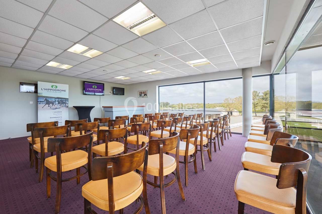Salle de conférence lumineuse avec rangées de chaises en bois, grandes baies vitrées et vue sur un paysage extérieur.