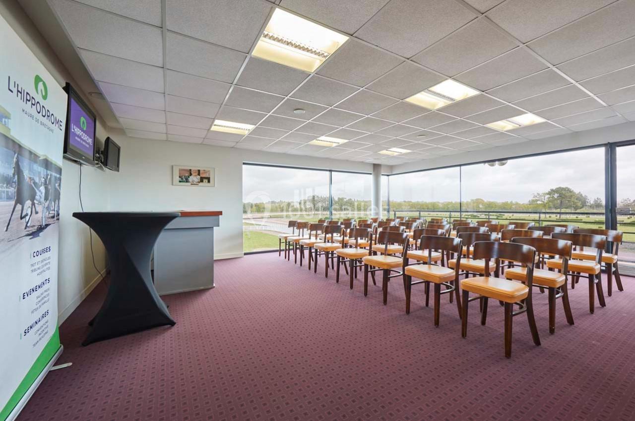 Salle de conférence lumineuse avec chaises alignées, pupitre, écran et vue sur un espace extérieur verdoyant.
