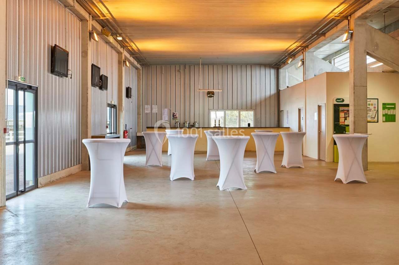 Salle lumineuse avec tables hautes blanches et bar en arrière-plan, murs en métal et sol en béton.