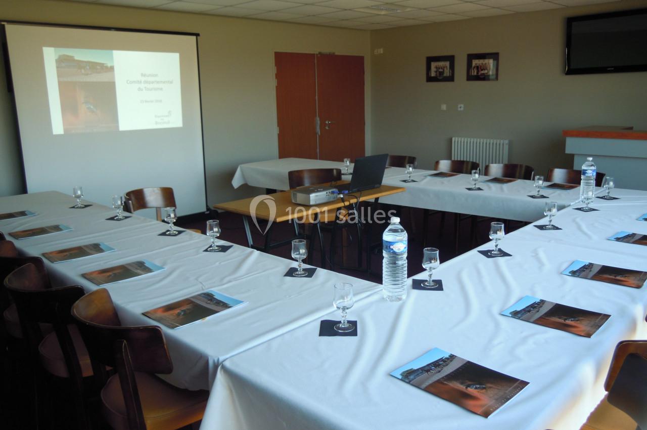 Salle de réunion aménagée en U avec des tables couvertes de nappes blanches, un projecteur et des documents disposés.