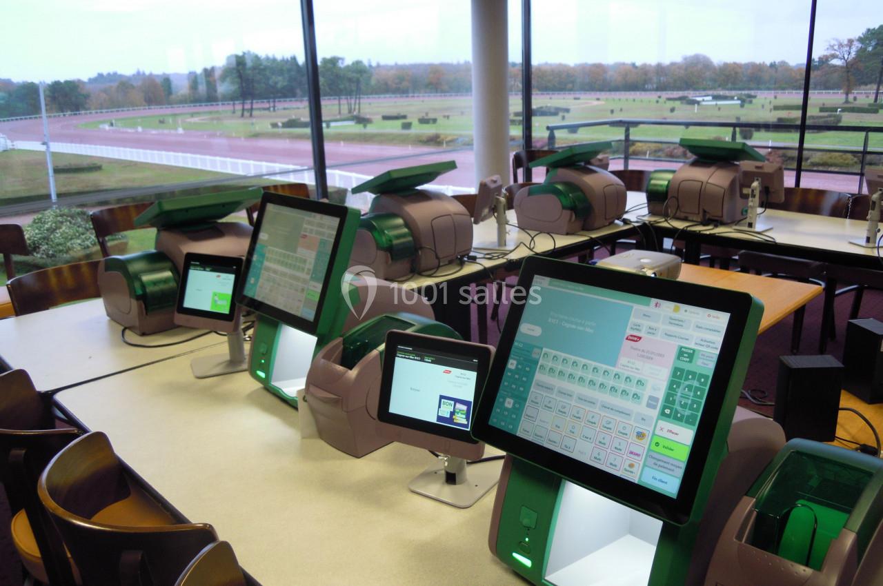 Des terminaux de paris électroniques alignés sur des tables dans une salle avec vue sur un hippodrome.