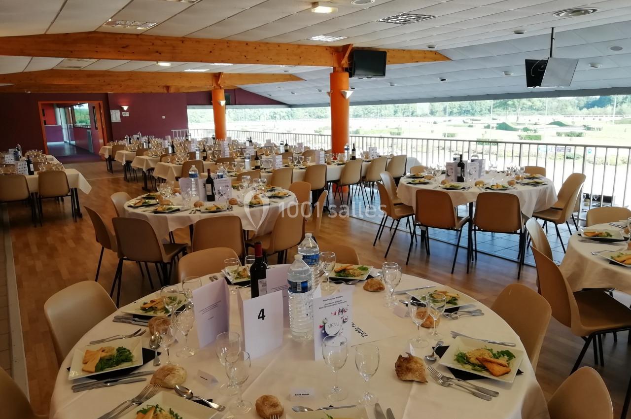 Salle de réception avec tables rondes dressées pour un repas, comprenant assiettes, verres, pain et entrées.
