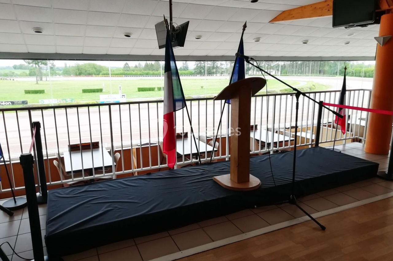 Estrade avec pupitre en bois, micros et drapeaux français, située dans une salle donnant sur un espace extérieur.