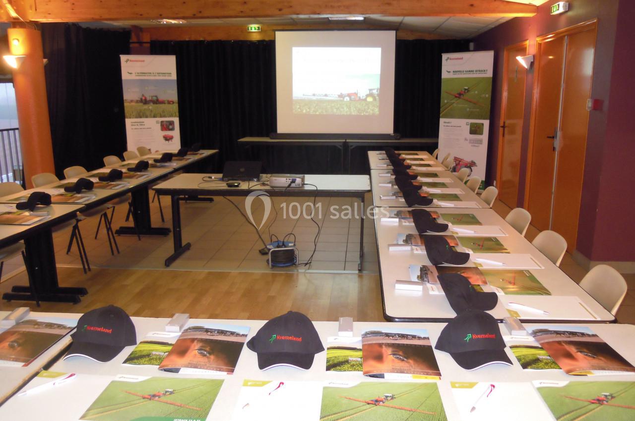 Salle de réunion avec tables disposées en U, documents, casquettes et écran de projection allumé en arrière-plan.