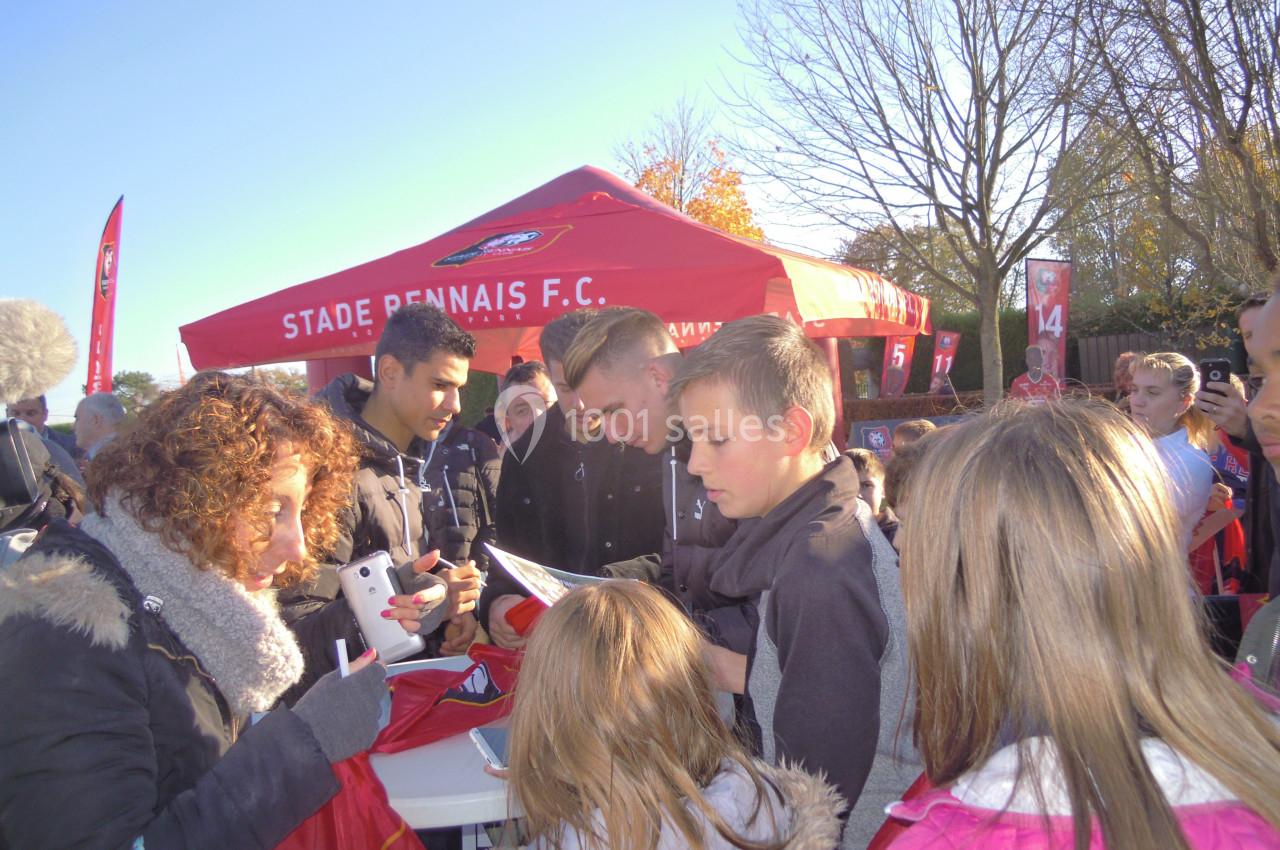 Des personnes rassemblées autour d'une table pour des autographes devant un stand du Stade Rennais F.C. en extérieur.