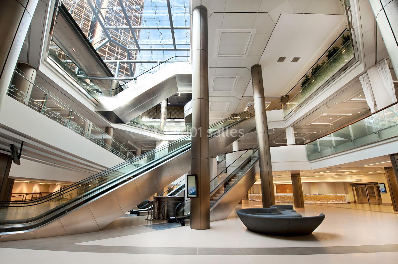 Intérieur moderne d'un bâtiment avec escalators, colonnes métalliques et verrière laissant entrer la lumière naturelle.