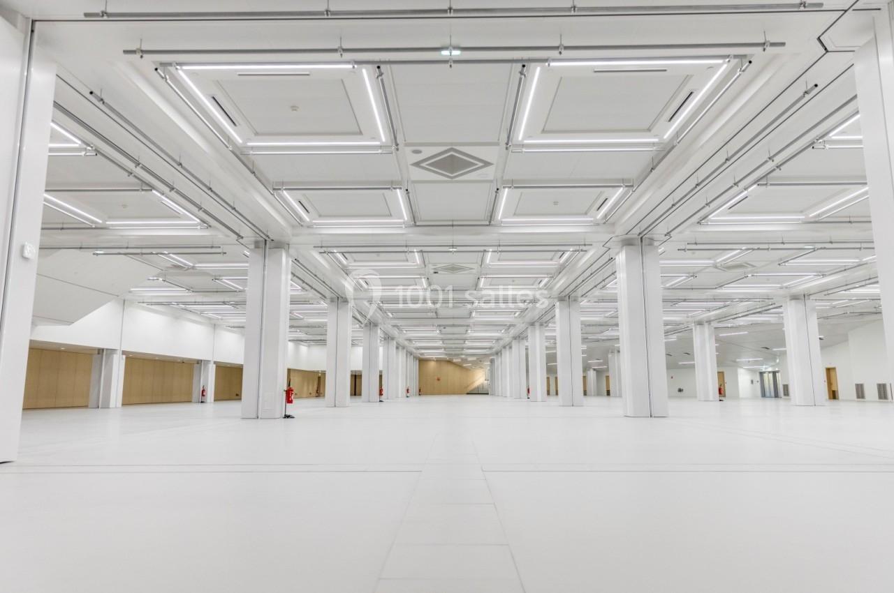 Grand espace intérieur vide avec un sol blanc, des colonnes et un plafond équipé de nombreux luminaires rectangulaires.