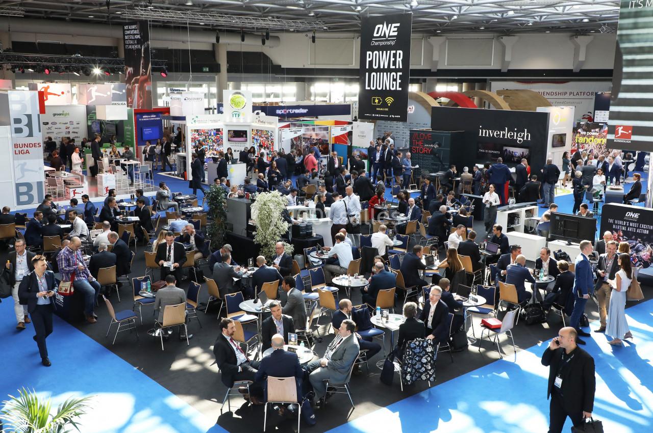 Salle d'exposition animée avec des stands, des visiteurs en discussion et des espaces de réunion.