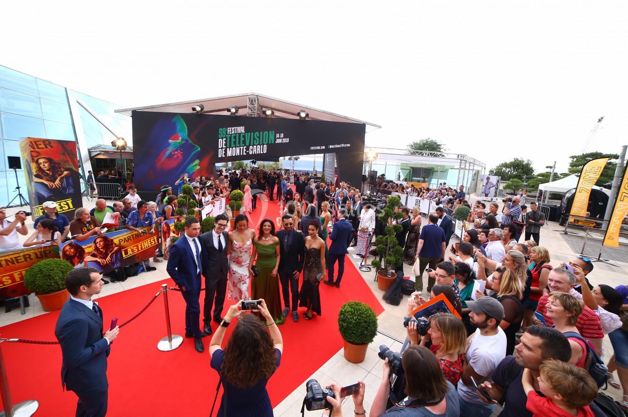 Des personnes posent sur un tapis rouge lors d'un événement en plein air, entourées de spectateurs et de photographes.