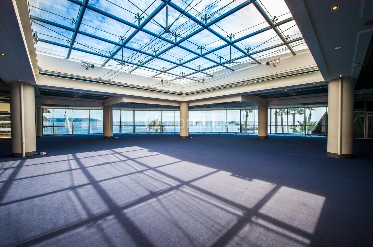 Salle spacieuse avec moquette bleue, grandes baies vitrées et plafond en verre laissant entrer la lumière naturelle.