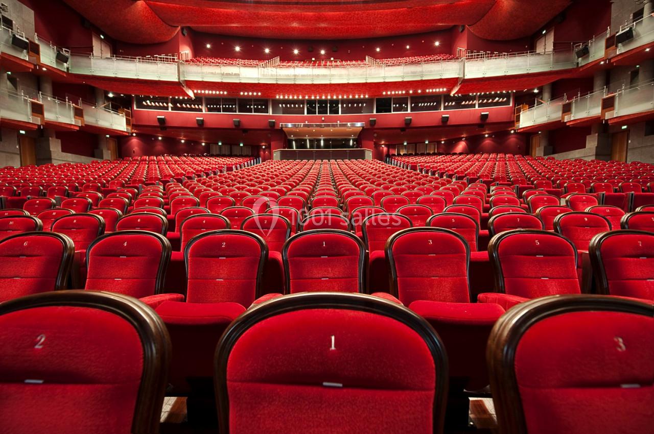 Salle de théâtre vide avec des rangées de sièges rouges alignés face à une scène éclairée.